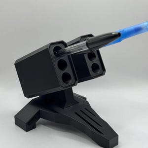May include: A black pen holder shaped like a missile launcher. The holder has multiple openings for pens and a sturdy base. A black pen and a blue pen are inserted into the openings, suggesting the product's function.