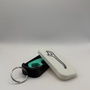 May include: A black and white keychain case with a silver key ring. The case is open, revealing a teal-colored dental floss pick and a white lid with a black floss pick outline.