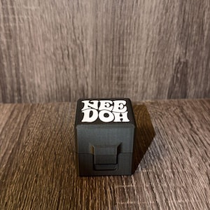 May include: A matte black square container with a hinged lid and a latch closure. The top of the lid features the white text "NEE DOH" printed in a bold, stylized font. The object has a smooth, solid construction with clean, sharp edges.