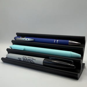 Op de afbeelding: Een zwarte pennenhouder met drie sleuven, met een blauwe pen, een lichtblauwe pen en een grijze Sharpie-marker. De pennenhouder is gemaakt van een materiaal met textuur en staat op een wit oppervlak.