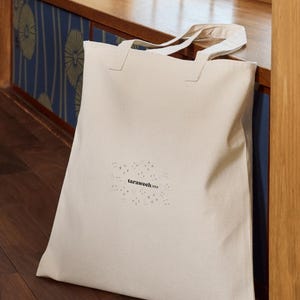 Puede incluir: Una bolsa de lona beige con la palabra "taraweeh" y gráficos de estrellas impresos en la parte delantera. La bolsa tiene dos asas y está apoyada contra una superficie de madera.