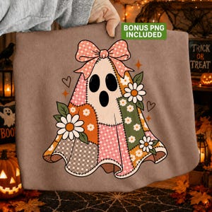 May include: A brown sweatshirt featuring a graphic of a ghost covered in a patchwork pattern of pink, orange, and green floral and checkered fabrics. The ghost is topped with a large pink checkered bow and decorated with white daisy flowers.