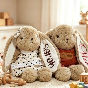 May include: Two plush tan bunnies with long ears. The left bunny wears a white dress with black polka dots and a lace trim. The right bunny wears rust-colored overalls with a brown patch. The name Sarah is embroidered on one ear, and Chloe on another.
