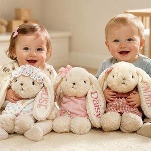 May include: Three cream-colored plush bunny toys with long floppy ears, each wearing a different outfit. The bunnies have names embroidered on their ears: Rose, Daisy, and Emma. They feature stitched facial details and soft, textured fur.