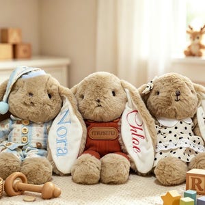 May include: Three plush tan bunnies with long ears. The left bunny wears blue plaid pajamas and a matching nightcap. The center bunny wears a rust-colored ribbed outfit with a brown patch. The right bunny wears a white dress with black polka dots.