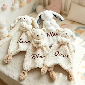 May include: Three plush security blankets featuring stuffed animal heads and textured fabric. One white rabbit with striped ears, one cream bear, and one cream lamb. Each blanket is embroidered with a name: Luna, Ethan, and Oscar in brown.