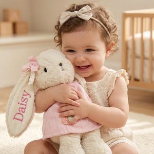 May include: A cream-colored plush rabbit toy featuring long ears, one of which is embroidered with the name Daisy in pink thread. The rabbit wears a light pink dress with a small floral detail and has a small pink ribbon bow attached to its ear.