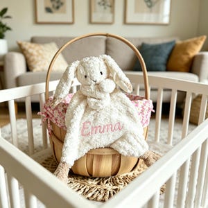 Puede incluir: Una manta de peluche con forma de conejito blanco con el nombre "Emma" bordado en rosa, dentro de una cesta de madera. La cesta está forrada con una tela floral y descansa sobre una esterilla tejida. El conejito tiene orejas largas y detalles en beige.