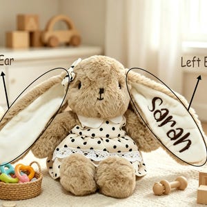 May include: A plush tan rabbit toy with long ears, wearing a cream-colored dress with black polka dots and a white lace trim. The left ear features the embroidered name Sarah in dark brown script, and a small black and white bow sits on the head.