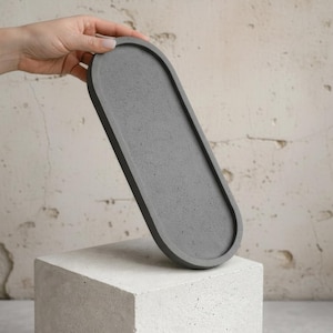 Pode incluir: Uma bandeja de concreto cinza em forma oval. A bandeja é segurada por uma mão e repousa sobre um bloco de concreto branco. A bandeja tem aproximadamente 30 cm de comprimento e 13 cm de largura, com uma borda elevada.