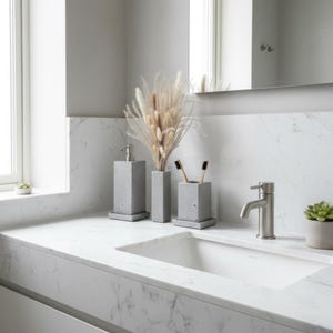 Puede incluir: Un conjunto de tres accesorios de baño de hormigón gris que incluye un dispensador de jabón, un jarrón alto y un soporte para cepillos de dientes, dispuestos sobre una encimera de mármol blanco junto a un grifo plateado y dos pequeñas suculentas.
