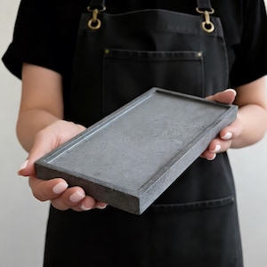Peut inclure: Un plateau rectangulaire en béton gris est présenté devant une personne portant un tablier noir. Le plateau a un design simple et minimaliste avec un bord légèrement surélevé. Le béton a une surface texturée et le plateau semble neuf.