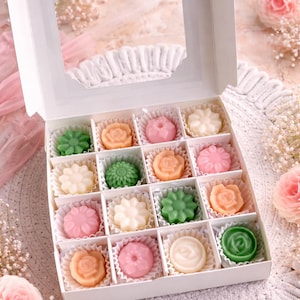 Puede incluir: Una caja blanca con una ventana transparente, llena de dieciséis jabones con forma de flor en varios colores pastel. Los jabones están dispuestos en copas de papel individuales. El fondo incluye flores decorativas y un tapete de encaje blanco.