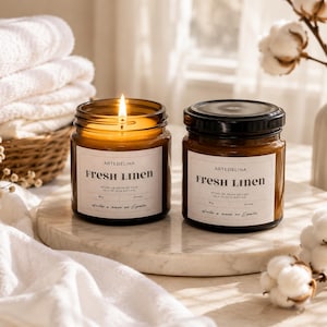 Pode incluir: Dois frascos de vidro âmbar contendo velas com rótulos brancos com a inscrição Artedelina Fresh Linen. Uma vela está acesa com uma chama visível, enquanto a outra está fechada com uma tampa preta. Os rótulos indicam um peso de 180g.