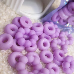 Pode incluir: Derretimentos de cera de cor lavanda em forma de donuts e esferas conectadas est&atilde;o espalhados sobre uma superf&iacute;cie branca texturizada. Um aquecedor de cer&acirc;mica branco est&aacute; no fundo, e um saco de derretimentos parcialmente vis&iacute;vel est&aacute; &agrave; direita.