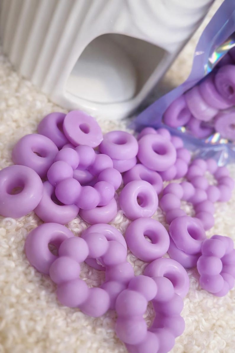 Pode incluir: Derretimentos de cera de cor lavanda em forma de donuts e esferas conectadas est&atilde;o espalhados sobre uma superf&iacute;cie branca texturizada. Um aquecedor de cer&acirc;mica branco est&aacute; no fundo, e um saco de derretimentos parcialmente vis&iacute;vel est&aacute; &agrave; direita.