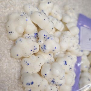 Pode incluir: Uma pilha de ceras perfumadas brancas em forma de nuvem com detalhes em glitter azul. As ceras est&atilde;o agrupadas, sugerindo um produto a granel. O fundo &eacute; um tecido macio e branco.