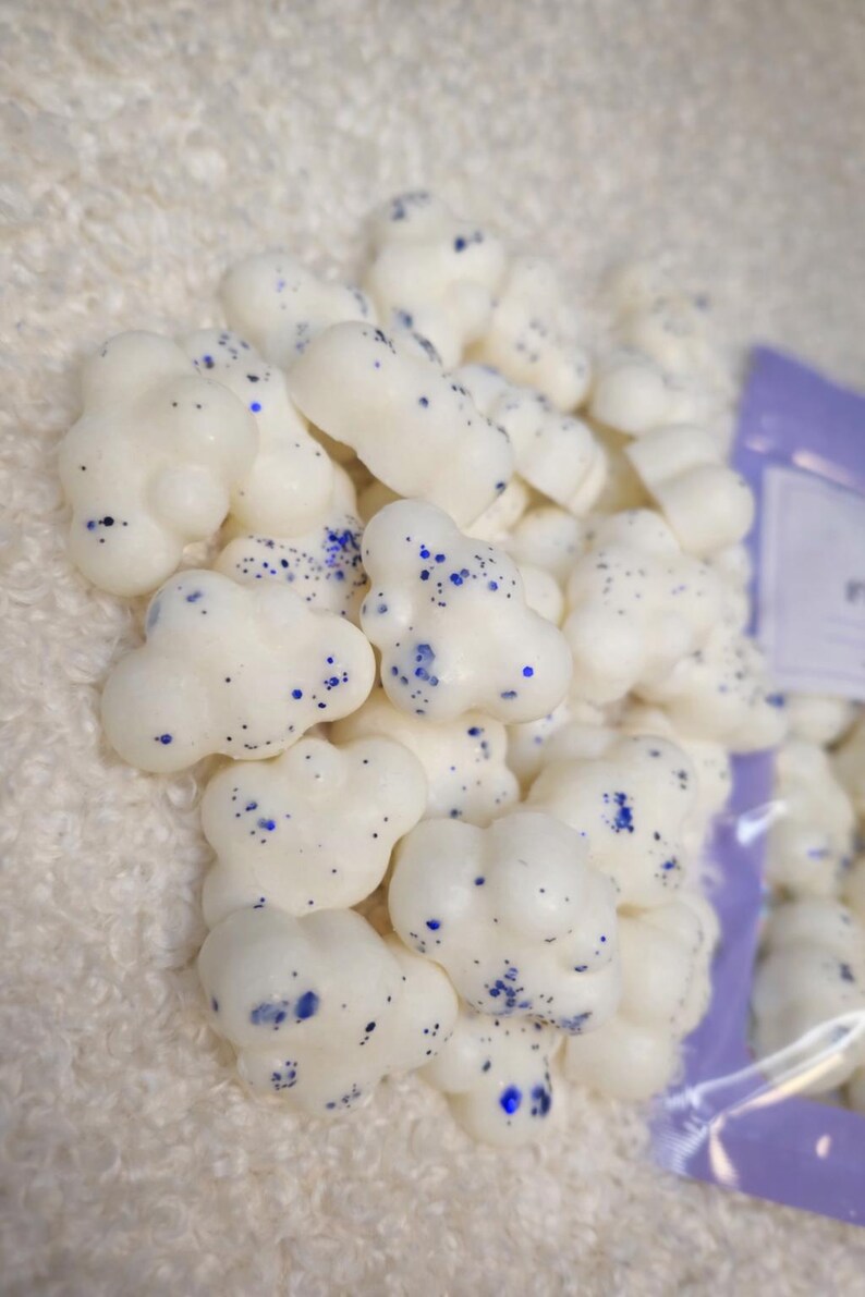 Pode incluir: Uma pilha de ceras perfumadas brancas em forma de nuvem com detalhes em glitter azul. As ceras est&atilde;o agrupadas, sugerindo um produto a granel. O fundo &eacute; um tecido macio e branco.