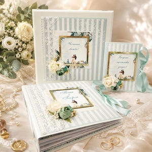 Puede incluir: Dos álbumes de fotos cuadrados y un libro de visitas rectangular con rayas verticales azul claro y blancas, paneles centrales con marco dorado con ilustraciones de una pareja y flores de tela decorativas con encaje y cintas azul claro.