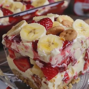 May include: A layered dessert featuring sliced bananas, strawberries, and cookies. The dessert has a creamy white filling and a red, jelly-like topping. The dessert is served in a clear glass dish, with a full pan of the dessert in the background.