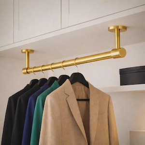 Puede incluir: Una barra de armario de metal dorado montada en el techo, con una barra horizontal sostenida por dos soportes circulares. Cinco perchas de plástico negro cuelgan de la barra, sosteniendo un blazer beige, uno verde, uno azul y dos blazers negros.
