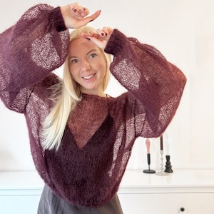 Può includere: Un maglione bordeaux a trama aperta con maniche lunghe e voluminose. Il maglione ha uno scollo tondo e un aspetto leggermente trasparente. Il design presenta una forma a diamante più scura e di colore pieno sul petto. Le maniche sono raccolte al polso.