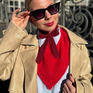 May include: A red knitted ascot scarf with a bow detail. The scarf is worn over a white and blue striped shirt and a red sweater. The person is wearing black sunglasses and a tan trench coat.
