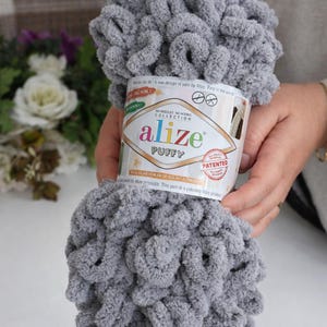 May include: A skein of gray Alize Puffy yarn. The yarn is held in a person's hands. The label reads "Alize Puffy" and "No Needles No Hooks Collection". The yarn is a soft, looped texture, ideal for various crafting projects.