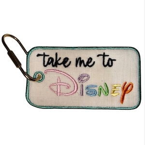 Puede incluir: Un llavero rectangular de tela color hueso con un borde bordado en color verde azulado y un cierre de metal dorado. El texto "take me to" está bordado en negro y la palabra "Disney" está bordada con hilo multicolor en rosa, azul, verde, amarillo y naranja.