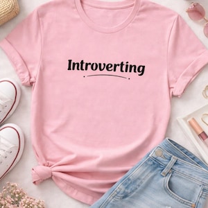 Könnte beinhalten: Ein hellrosa T-Shirt mit dem Wort "Introverting" in schwarzer Schrift. Das Shirt wird mit Jeansshorts, weißen Turnschuhen und Accessoires kombiniert. Das T-Shirt ist an der Taille geknotet.