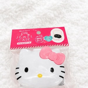 以下が含まれることがあります： 透明なビニール袋に入った白いハローキティのジュエルケース。ケースはハローキティの顔の形をしており、ピンクのリボン、黒い目、ひげがあります。パッケージには日本語のテキストとピンクのアクセントが含まれています.