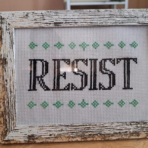 May include: Cross-stitch art in a distressed white wooden frame. The word "RESIST" is stitched in black thread on a white background, with green diamond-shaped accents above and below the text. The frame has a rustic, weathered appearance.