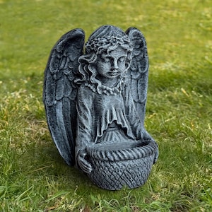May include: A gray angel statue with detailed wings and a basket-shaped container. The angel is seated, with a serene expression, and is surrounded by green grass. The statue is made of a stone-like material.