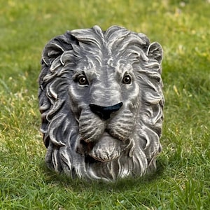 May include: A detailed, gray lion head sculpture with a black nose and detailed mane. The sculpture is set against a backdrop of green grass, suggesting an outdoor garden decor item.