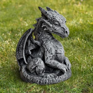 May include: A detailed, gray dragon statue with textured scales, wings, and a stern expression. The mythical creature is seated on a circular base, set against a backdrop of green grass. The statue is a decorative garden ornament.