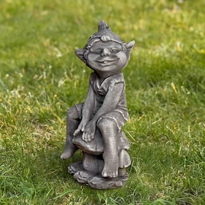 May include: A gray garden gnome statue sitting on a mushroom. The gnome has a smiling face, pointed ears, and is wearing a hat and tunic. The statue is made of a stone-like material and is placed on a grassy lawn.