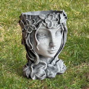 May include: A gray and white planter shaped like a woman's face, adorned with a floral crown and flowing hair. The detailed sculpture is made of a textured material and sits on a bed of green grass. A decorative item for the garden.