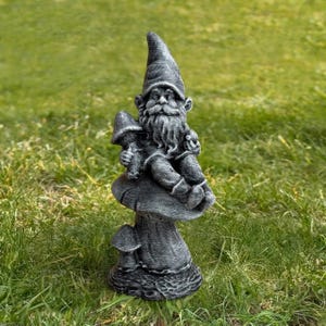 May include: A gray garden gnome statue with a long beard and pointed hat, holding a mushroom. The gnome is seated on a large mushroom, with smaller mushrooms at the base. The statue is made of a stone-like material.
