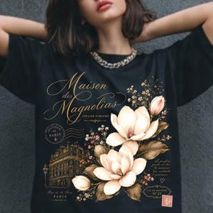 May include: A black t-shirt featuring a graphic print with the text Maison des Magnolias Atelier Parfumé. The design includes illustrations of large cream-colored magnolia flowers, brown leaves, a Parisian building, and a small pink postage stamp.