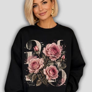 Peut inclure: Un sweat-shirt noir à col rond arborant un grand imprimé graphique représentant trois roses roses en fleurs avec des feuilles vertes. Le motif floral est superposé sur un fond de texte style journal vieilli blanc cassé incluant 1983.
