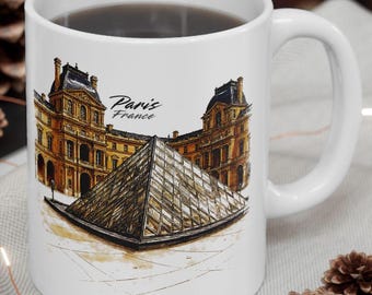 May include: White ceramic mug featuring a detailed illustration of the Louvre Museum and the glass pyramid in Paris, France. The mug has a curved handle and the words "Paris France" are printed on the front.