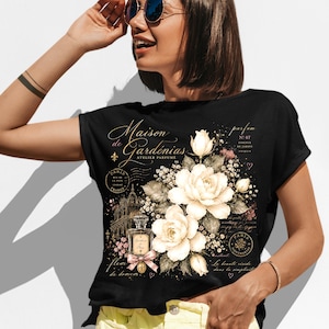 May include: A black t-shirt featuring a graphic print of white gardenia flowers, a perfume bottle with a pink bow, and French text that reads Maison de Gardénias, Atelier Parfumé, Paris, and other decorative script elements in gold and white.
