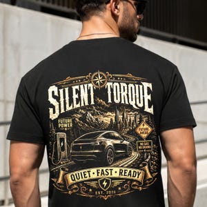 Op de afbeelding: Een zwart t-shirt met een afbeelding op de achterkant met de tekst SILENT TORQUE, FUTURE POWER, FUTURE AHEAD, NO GAS NO DRAMA en QUIET FAST READY. De illustratie toont een elektrische auto, een oplaadstation en een stadsskyline.