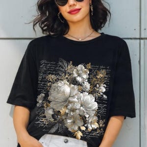 May include: A black short-sleeved t-shirt featuring a central graphic of silver and white floral blossoms layered over a background of black cursive script text. The design includes detailed petals and leaves in a monochromatic colour palette.