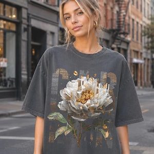 May include: A charcoal gray short-sleeve t-shirt featuring a large, central graphic of a white peony flower with gold-toned accents. The design incorporates a fragmented, glitch-style aesthetic with vertical lines and metallic gold leaf details.