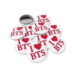 以下が含まれることがあります： 白い丸いバッジのコレクションで、それぞれに赤い文字で「I ♥ BTS」と赤いハートのシンボルが表示されています。1つのバッジはピンの裏側を示しています。バッジは白い背景に配置されています。