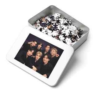 May include: A white puzzle box with a group photo of seven people in black clothing. The puzzle pieces are in a silver tin. The puzzle pieces are white with a purple and black design.