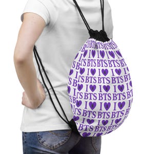 May include: White drawstring backpack with a repeating pattern of purple hearts and the text "I BTS". The bag has black drawstrings and a black closure. The bag is worn over the shoulder.