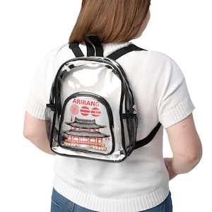 Puede incluir: Mochila transparente con ribete negro y bolsillos laterales de malla. La mochila presenta un gráfico de un edificio de estilo asiático rojo y negro con la palabra "ARIRANG" en texto rojo.
