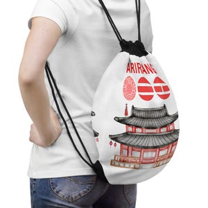 May include: White drawstring backpack featuring a red and black Asian-style building design. The word "ARIRANG" is printed in red at the top, along with other red graphic elements. The bag has black drawstrings.